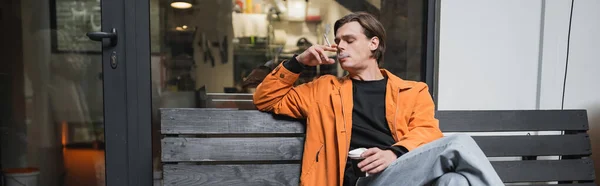 Stylish man holding takeaway drink and smoking drink on bench near cafe, banner — Stock Photo