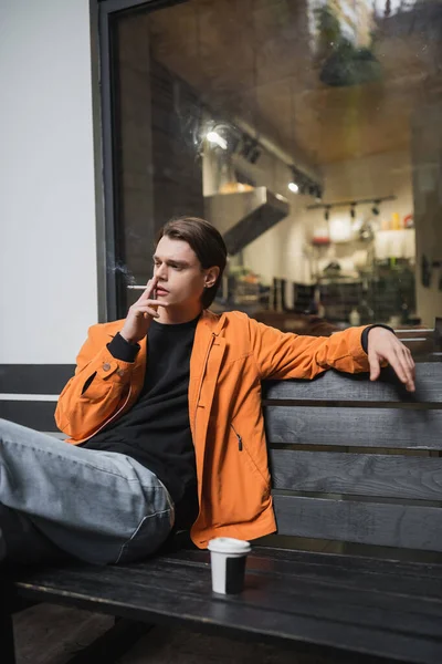 Trendy young man smoking cigarette near coffee to go on bench outdoors — Stock Photo