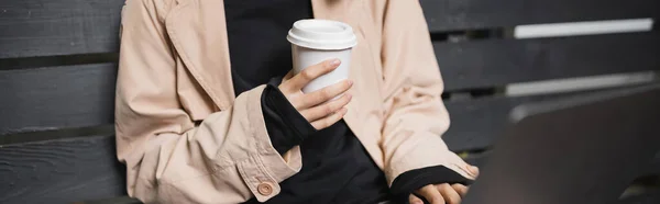 Vista cortada de jovem freelancer em casaco de trincheira segurando copo de papel e usando smartphone no banco, banner — Fotografia de Stock