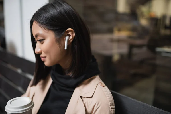 Vista lateral da jovem mulher asiática no fone de ouvido segurando copo de papel enquanto sentado no banco perto do café — Fotografia de Stock