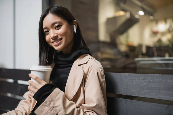 Sorrindo mulher asiática em casaco de trincheira segurando café para ir e olhando para a câmera no banco perto de café — Fotografia de Stock