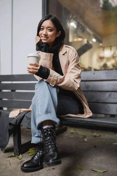 Stylish asian woman in trench coat holding paper cup and looking at camera on bench near cafe — Stock Photo