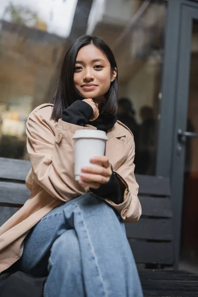 Moda mulher asiática em casaco de trincheira segurando copo de papel no banco perto de café — Fotografia de Stock
