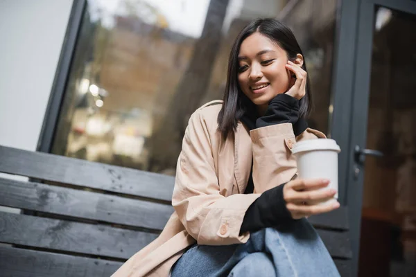 Alegre ásia mulher no trincheira casaco segurando takeaway café enquanto sentado no banco perto de café — Fotografia de Stock
