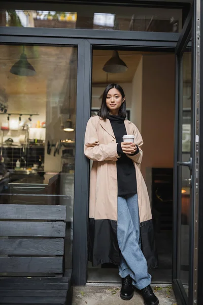 Модна азіатська жінка в траншеї пальто тримає паперовий стакан біля кафе — стокове фото