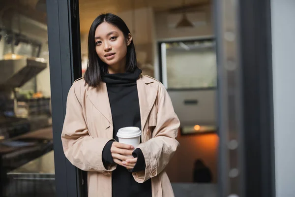 Mulher asiática bonita no casaco de trincheira que prende o café para ir perto da porta aberta do café — Fotografia de Stock