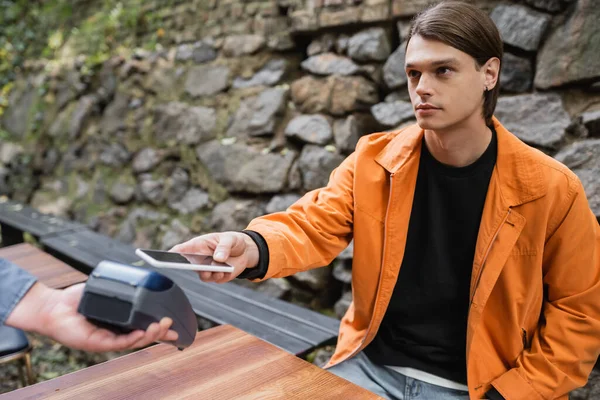 Camarera borrosa sosteniendo terminal de pago cerca del joven con teléfono celular en la terraza de la cafetería - foto de stock