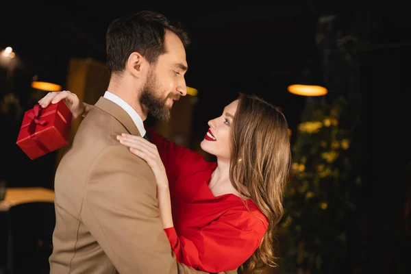 Side view of cheerful woman holding present and hugging bearded boyfriend on valentines day — Stock Photo