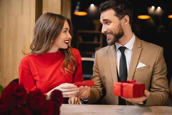 Homem alegre e barbudo dando cartão de saudação em forma de coração vermelho enquanto segurando presente perto da namorada feliz — Fotografia de Stock