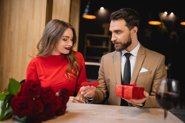 Barbudo hombre dando rojo en forma de corazón tarjeta de felicitación mientras sostiene presente cerca de novia feliz - foto de stock
