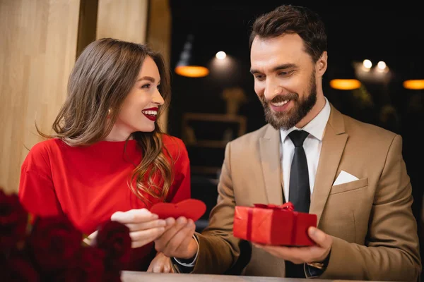 Alegre joven dando tarjeta de felicitación en forma de corazón rojo mientras sostiene presente cerca de novia feliz - foto de stock
