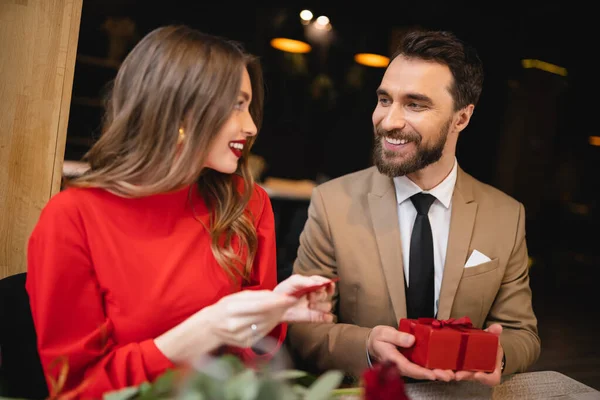 Hombre alegre y barbudo sosteniendo presente cerca de novia feliz con tarjeta de felicitación en forma de corazón rojo - foto de stock