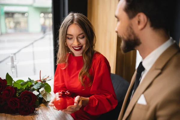 Femme gaie en robe rouge tenant cadeau en forme de coeur avec arc près de petit ami le jour de la Saint-Valentin — Photo de stock