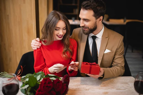 Cheerful man holding present near happy girlfriend reading heart-shaped greeting card — Stock Photo