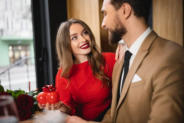 Mulher alegre em vestido vermelho segurando presente em forma de coração com arco e olhando para o namorado no dia dos namorados — Fotografia de Stock