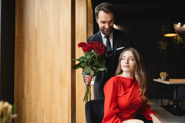 Bearded man in suit smiling while holding bouquet of red roses near girlfriend on valentines day — Stock Photo
