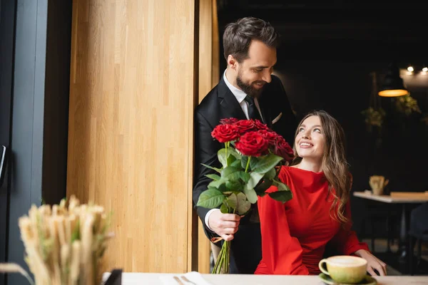 Barbudo homem no terno segurando buquê de rosas vermelhas perto de sorrir namorada no dia dos namorados — Fotografia de Stock