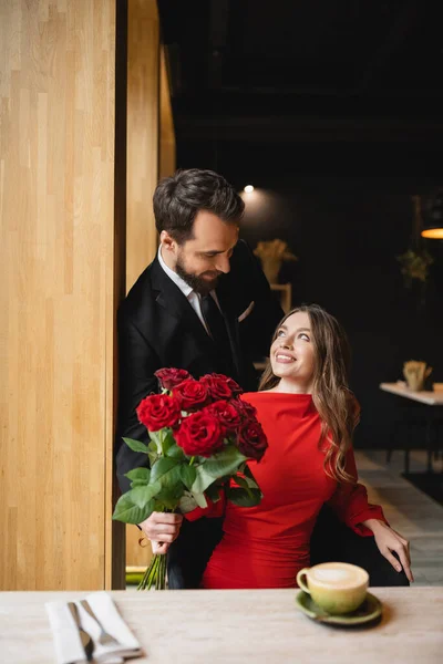 Hombre barbudo en traje con ramo de rosas rojas cerca de novia feliz en el día de San Valentín - foto de stock