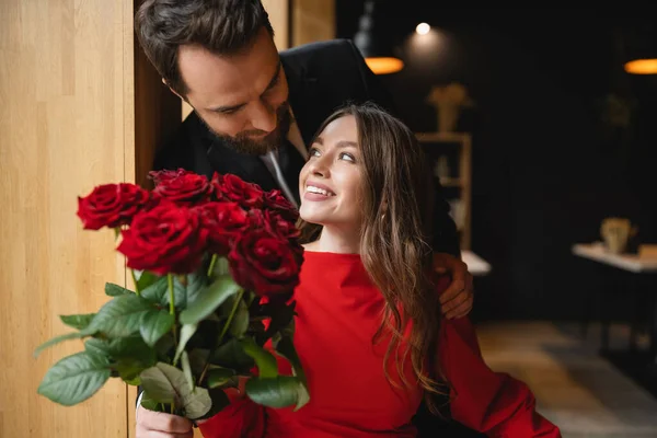 Uomo barbuto in vestito che tiene mazzo di rose rosse vicino a fidanzata gioiosa durante il giorno di San Valentino — Foto stock