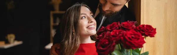 Homem barbudo segurando buquê de rosas vermelhas perto de namorada feliz no dia dos namorados, banner — Fotografia de Stock