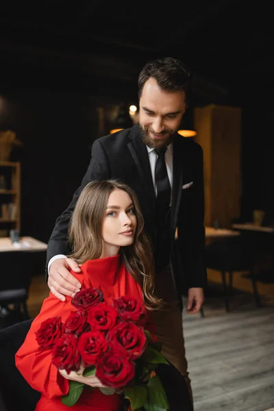 Hombre alegre en traje sonriendo mientras mira a la novia sosteniendo ramo de rosas rojas en el día de San Valentín - foto de stock