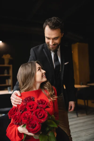 Bearded man in suit smiling while looking at happy girlfriend holding bouquet of red roses on valentines day — Stock Photo