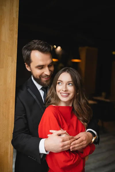 Hombre barbudo en traje abrazando a mujer feliz en vestido rojo en el día de San Valentín - foto de stock