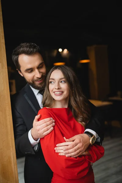 Barbudo homem no terno abraçando alegre mulher no vestido vermelho no dia dos namorados — Fotografia de Stock