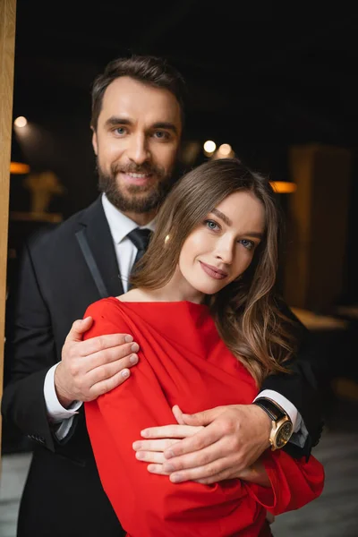 Hombre barbudo en traje abrazando mujer bonita y feliz en vestido rojo en el día de San Valentín - foto de stock