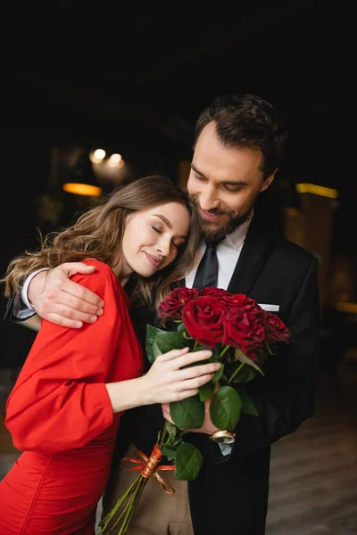 Barbudo hombre abrazando feliz novia con ramo de rosas rojas en día de San Valentín - foto de stock