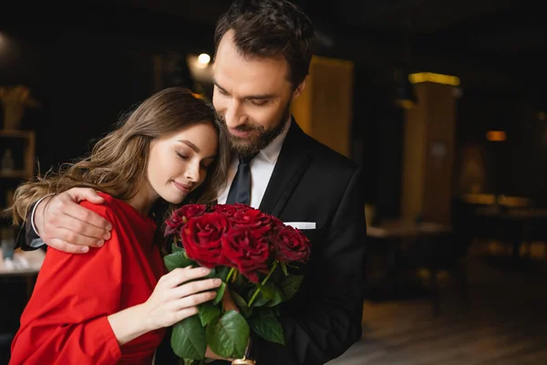 Bearded man hugging smiling girlfriend with bouquet of red roses on valentines day — Stock Photo