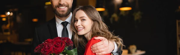 Barbudo hombre abrazando feliz novia con ramo de rosas rojas en día de San Valentín, bandera - foto de stock