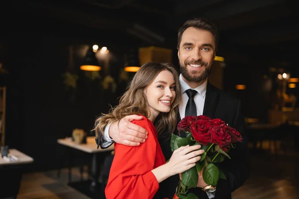 Smiling man hugging happy girlfriend near bouquet of red roses on valentines day — Stock Photo
