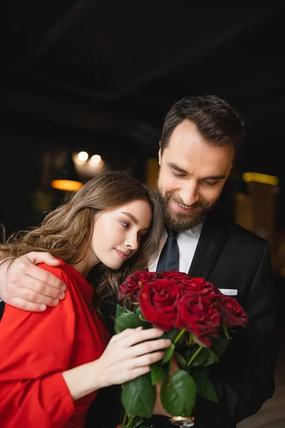 Pleased man hugging happy girlfriend with bouquet of red roses on valentines day — Stock Photo