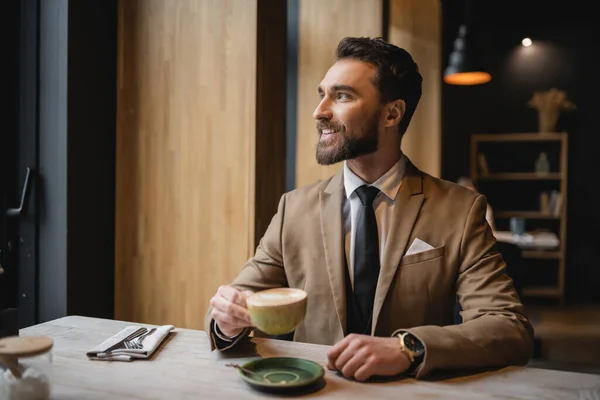 Felice uomo barbuto in giacca e cravatta in possesso di tazza di cappuccino in caffè — Foto stock