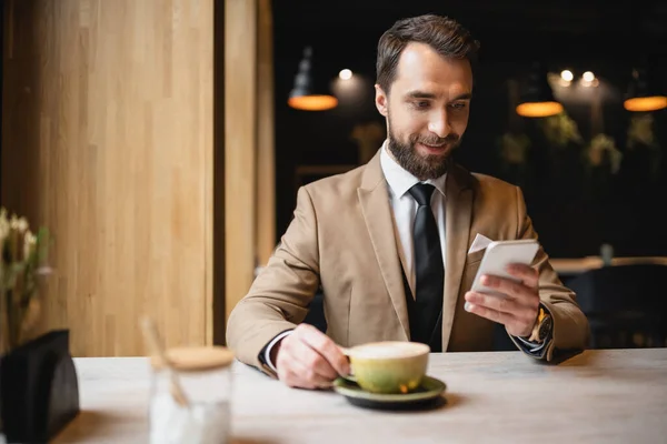 Cheerful man with beard using smartphone while holding cup of cappuccino in cafe — Stock Photo