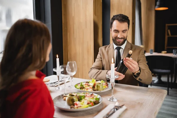 Bärtiger Mann lächelt, während er beim festlichen Abendessen am Valentinstag eine Flasche mit Wein in der Nähe seiner Freundin hält — Stockfoto