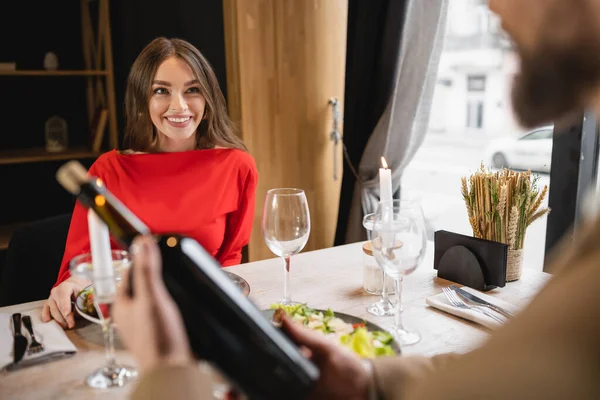 Fröhliche Frau im roten Kleid schaut Mann an, der am Valentinstag Flasche mit Wein hält — Stockfoto