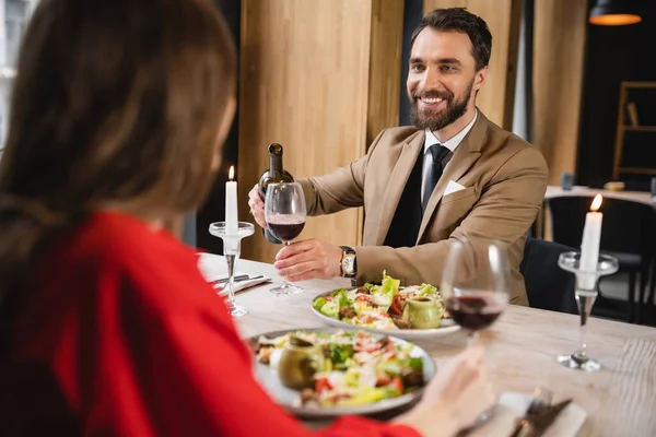 Uomo allegro versando vino rosso in vetro vicino fidanzata durante la cena il giorno di San Valentino — Foto stock