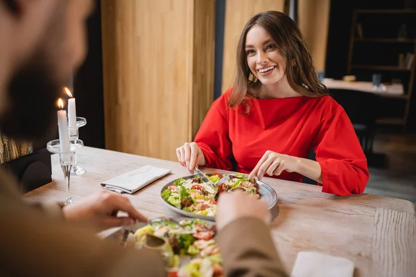 Fröhliche Frau lächelt und schaut ihren Freund an, während sie am Valentinstag Besteck in der Nähe von Salat im Restaurant hält — Stockfoto