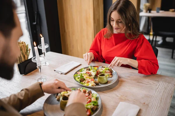 Cheerful woman smiling and looking at salad near man in restaurant on valentines day — Stock Photo