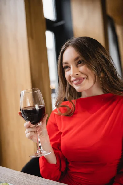 Feliz joven mujer sosteniendo copa con vino tinto y sonriendo en el restaurante - foto de stock