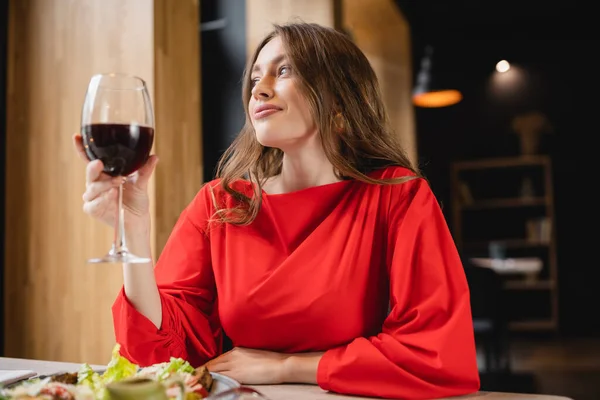Feliz joven mujer sosteniendo vaso con vino tinto y sonriendo cerca del plato con ensalada en el restaurante - foto de stock