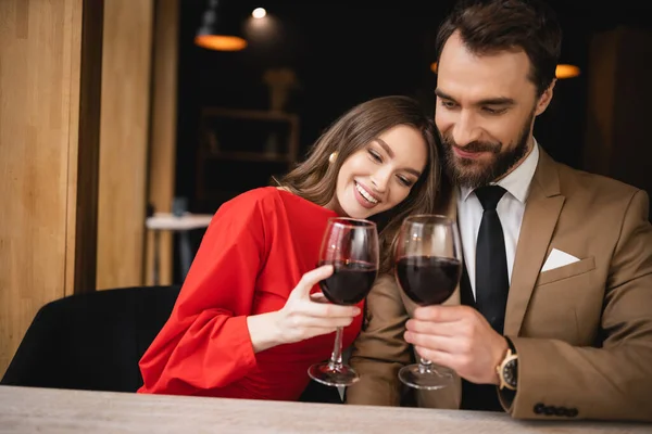 Mulher alegre e barbudo homem clinking copos com vinho tinto durante a celebração no dia dos namorados — Fotografia de Stock