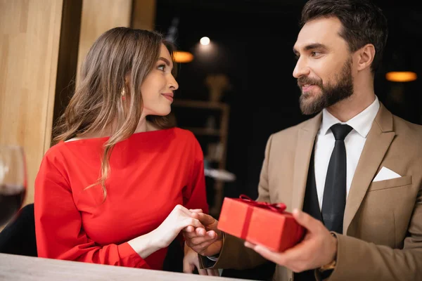 Бородатий чоловік в офіційному одязі тримає подарунок біля усміхненої жінки в червоній сукні в день валентинки — стокове фото