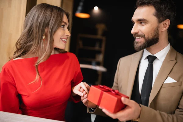 Homem barbudo no desgaste formal segurando presente perto mulher feliz no vestido vermelho no dia dos namorados — Fotografia de Stock