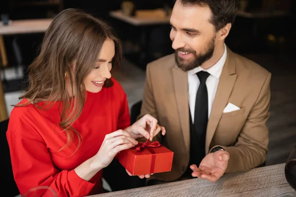 Barbudo hombre en formal desgaste celebración presente mientras que la mujer feliz en vestido rojo tirando de la cinta en el día de San Valentín - foto de stock