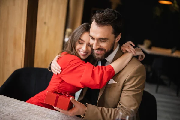 Cheerful woman in red dress hugging bearded man in formal wear holding present on valentines day — Stock Photo