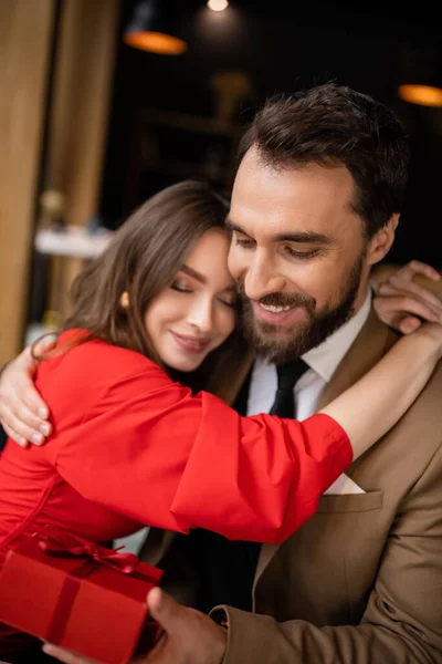 Happy woman in red dress hugging bearded boyfriend in formal wear holding present on valentines day — Stock Photo