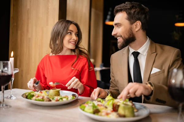 Jovem casal alegre em trajes festivos olhando um para o outro, tendo refeição durante a celebração no dia dos namorados — Fotografia de Stock